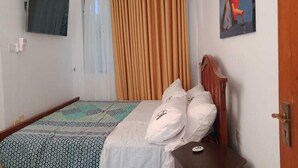 A bedroom with a bed, a wooden headboard, a bedside table, a wall-mounted TV, a curtain, and a painting. - Lux Suites Royal Palm Studio Apartment (Mtwapa)