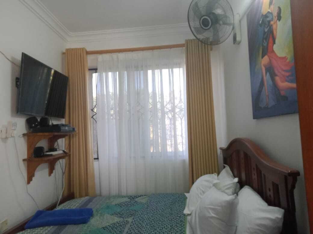 A bedroom with a bed, a TV on a shelf, a fan, and a painting on the wall.