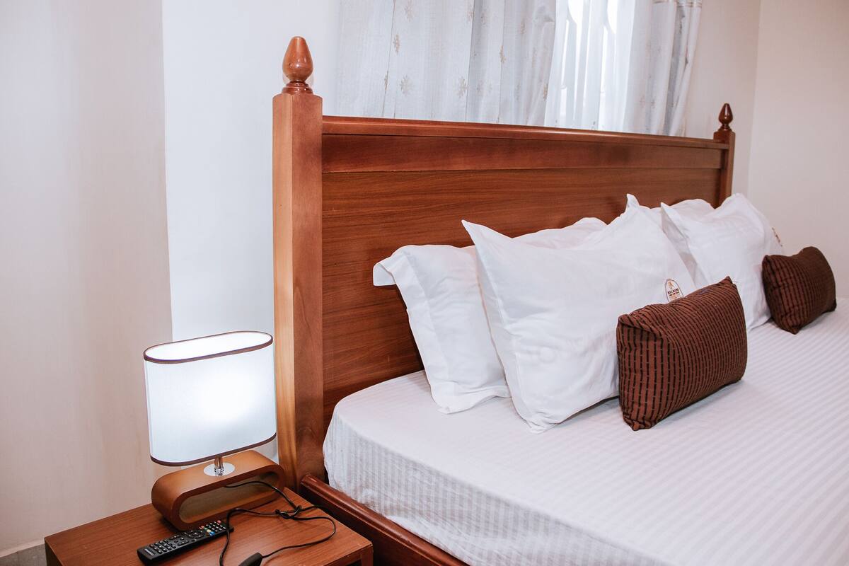 Ein ordentlich bezogenes Bett mit weißen und braunen Kissen, ein Holz-Kopfteil, eine Nachttischlampe und eine Fernbedienung auf einem Nachttisch aus Holz.