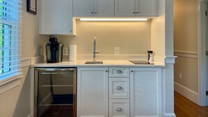 A compact kitchen with white cabinetry, a built-in dishwasher, and a countertop with a coffee maker and a kettle. - West End - Commercial Street, Charming 2-Bedroom with Beach Access - Sleeps 4! (Provincetown)
