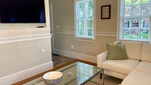 A living room with a flat-screen TV, a glass coffee table, a white sofa, and two windows with blinds. - West End - Commercial Street, Charming 2-Bedroom with Beach Access - Sleeps 4! (Provincetown)