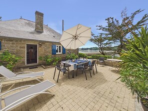 Ein Steinhaus mit einer gefliesten Terrasse, Sitzgelegenheiten im Freien und Blick auf das Wasser.