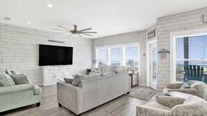 A spacious living room with a large flat-screen TV, a ceiling fan, and a view through the glass doors. - Gulf Front Luxury | Private Pool and Elevator (Gulf Shores)