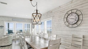 A dining area with a wooden table, white chairs, and a large wall clock. - Gulf Front Luxury | Private Pool and Elevator (Gulf Shores)