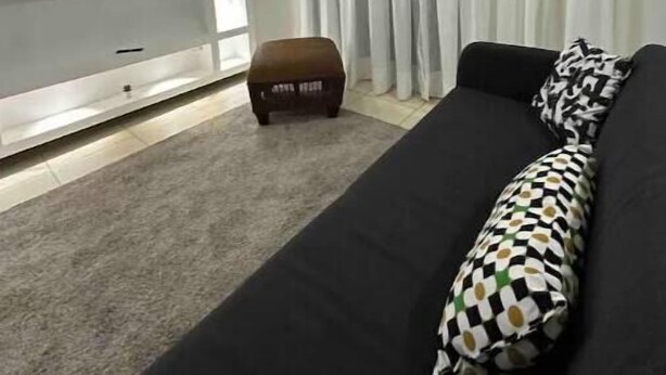 A modern living room with a black sofa, patterned throw pillow, and a flat-screen TV mounted on a white wall. - Burungos Place @ Terrazas Del Caribe D-202 (Santo Domingo Este)
