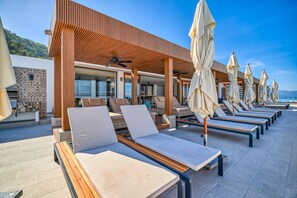 A sunlit outdoor lounge area with rows of lounge chairs and umbrellas, a wooden pergola, and a view of the sea. - Amapas Oasis | Pool & Private BBQ (Puerto Vallarta)