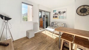 A modern living space with a wooden floor, a dining area with a wooden table and benches, a sofa with a geometric-patterned cover, a large window, and a kitchen visible through a glass door.