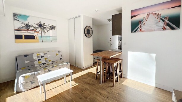 A compact living space with a sofa, a dining table with stools, a kitchen area, and artwork on the walls.