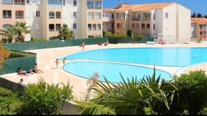 A swimming pool surrounded by multi-story buildings with balconies, a clear blue sky, and some greenery.