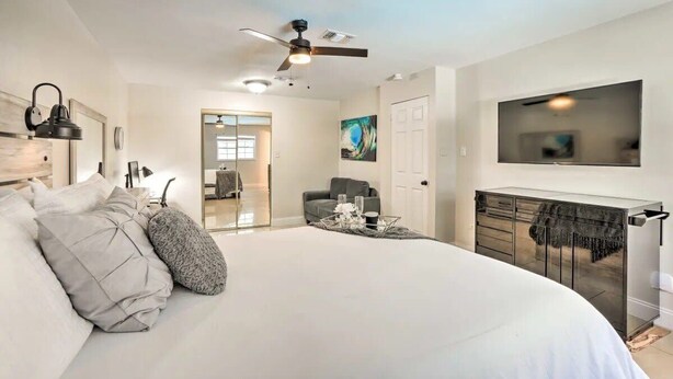 A modern bedroom with a large bed, a ceiling fan, a TV mounted on the wall, and a glass door leading to another room. - SunRay Stay ~ Heated Pool, Putting Green, & Patio (Safety Harbor)