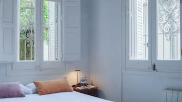 A bedroom with a bed, pillows, a nightstand with a lamp, and two windows with shutters.