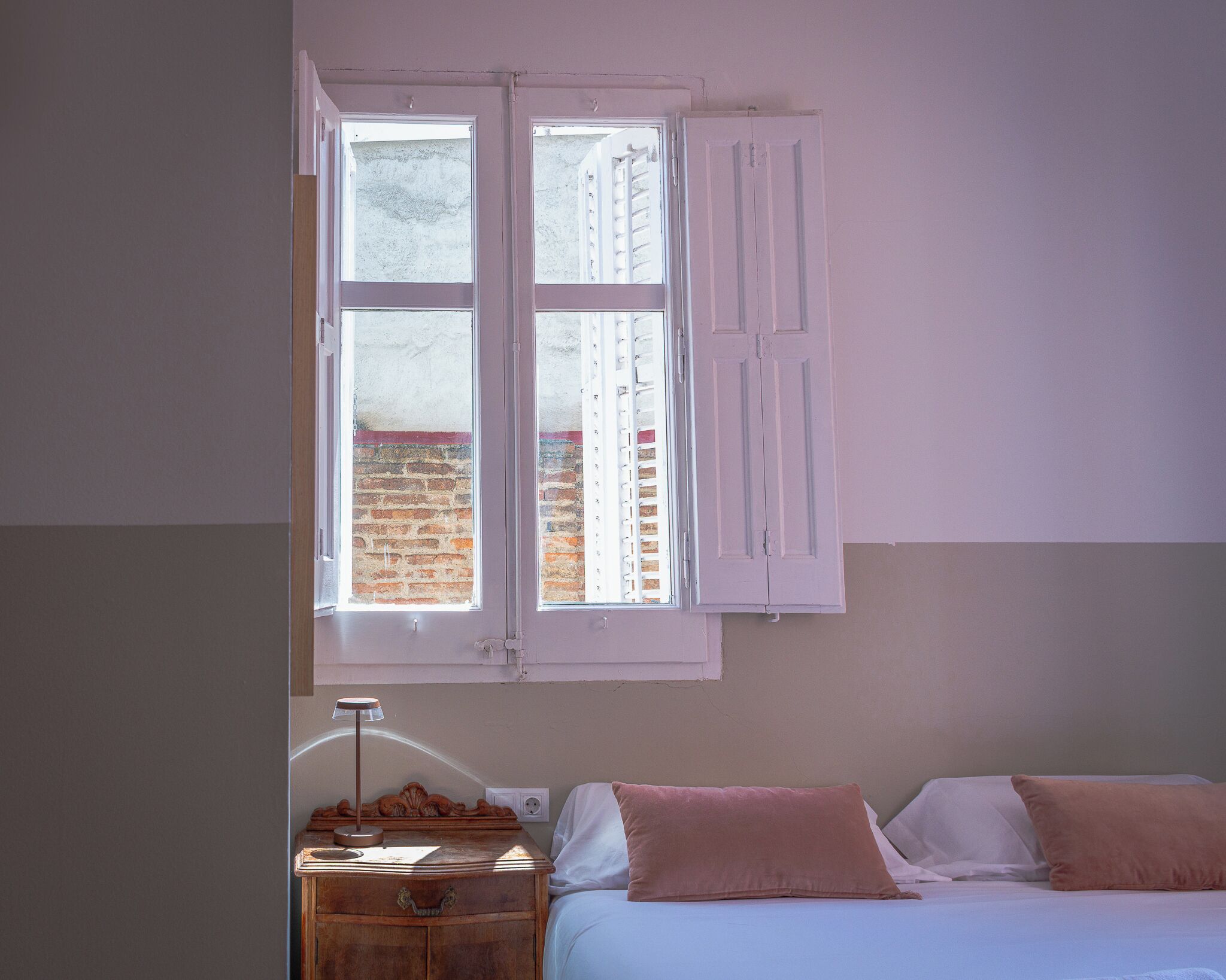 Un dormitorio con cama, mesita de noche, lámpara y ventana con contraventanas.