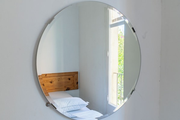A round mirror reflecting a bed with a wooden headboard, a window, and a view of greenery outside. - Verdi 254 (Barcelona)