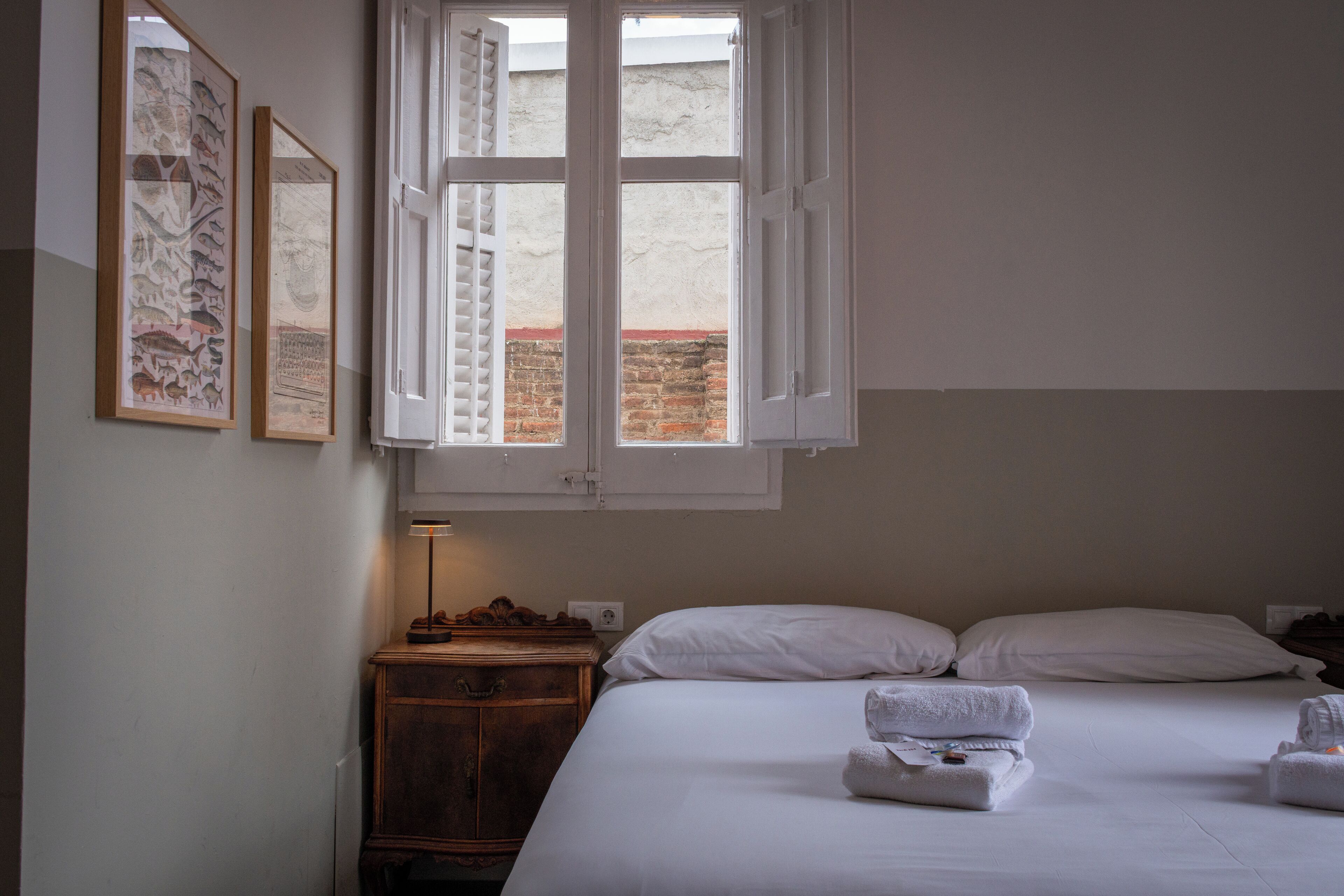 Hotellihuone, jossa on sänky, valkoiset vuodevaatteet, puinen yöpöytä, lamppu ja kaksi kehystettyä kuvaa seinällä.