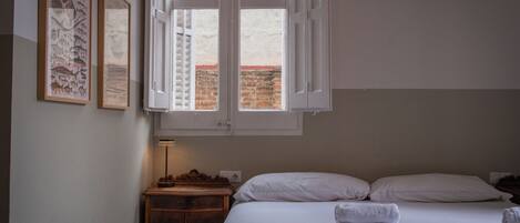 Hotellihuone, jossa on sänky, valkoiset vuodevaatteet, puinen yöpöytä, lamppu ja kaksi kehystettyä kuvaa seinällä.