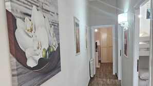 En hall med trägolv, vita väggar och en stor tavla med blommotiv. Här finns dörrar som leder till olika rum, ett element och en lampa.