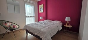 A bedroom with a bed, a chair, two bedside tables with lamps, a window with curtains, and a wall with a painting.