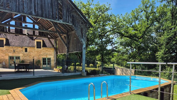 Ein Schwimmbad mit Holzterrasse, ein Steinhaus und ein eingezäunter Bereich mit Gartenmöbeln.