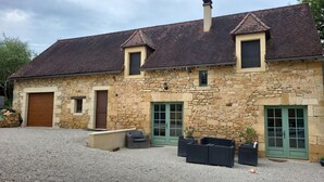 Ein Steinhaus mit Ziegeldach, zwei Fenster mit grünen Rollläden und eine Tür. Es gibt Außenbereiche mit Sitzgelegenheiten und Pflanzgefäßen.