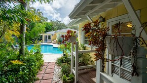 Un espace piscine doté d’une terrasse en bois, entouré d’une végétation luxuriante et bordé d’une maison à la façade jaune.