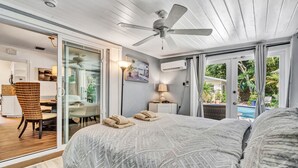 Une chambre avec un lit, un ventilateur de plafond, un bureau avec une chaise, une lampe et une vue sur une piscine et la verdure environnante.