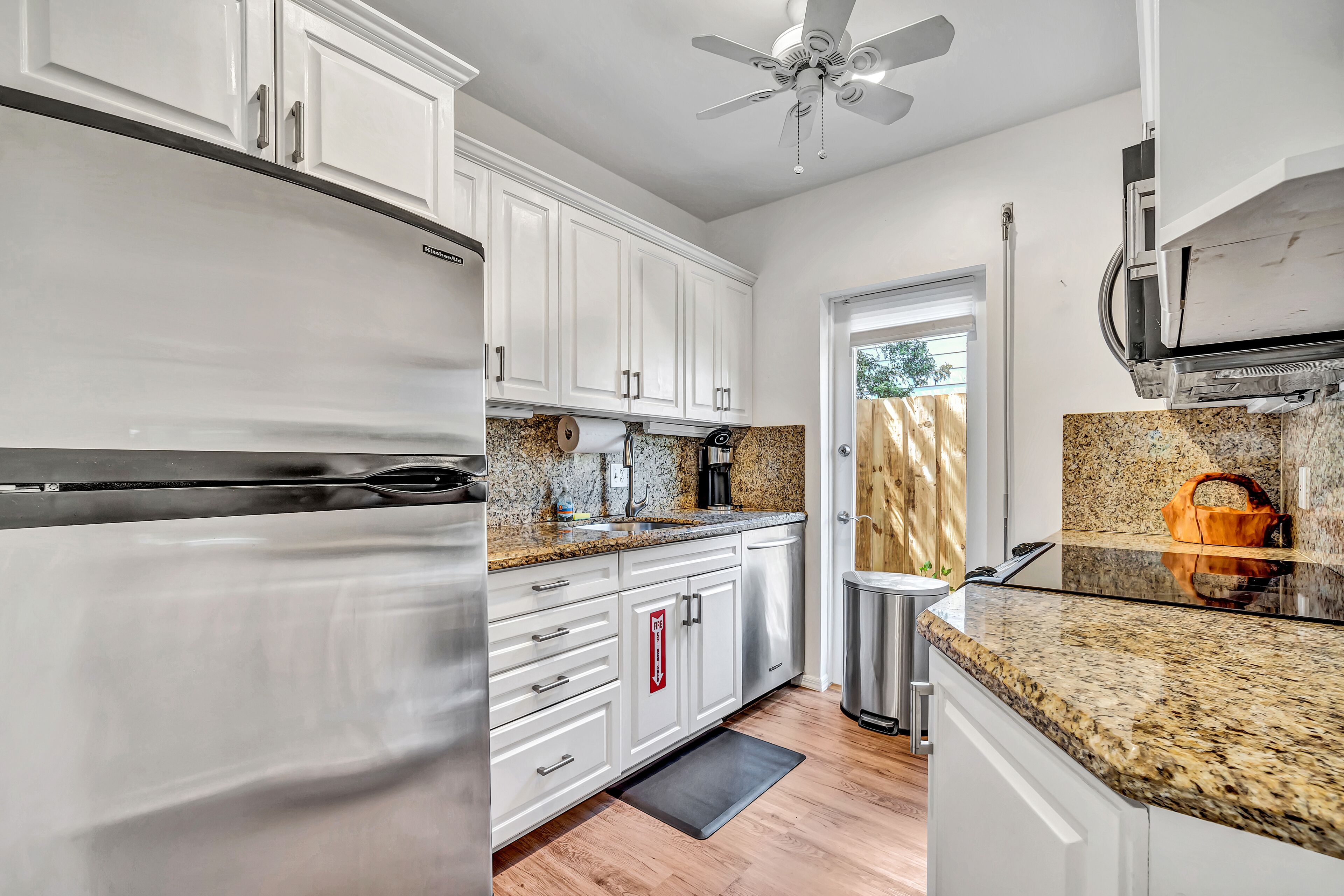 A modern kitchen with white cabinets, granite countertops, a stainless steel refrigerator, and a ceiling fan.