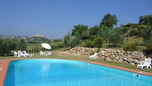 Une piscine entourée de verdure et d’un mur de pierre, avec des chaises longues blanches et une table agrémentée d’un parasol.