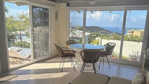 A sunlit room with a dining table and chairs, a sofa, and a view of the ocean. - Seabreeze Cottage with private  pool and sunset view (Cruz Bay)