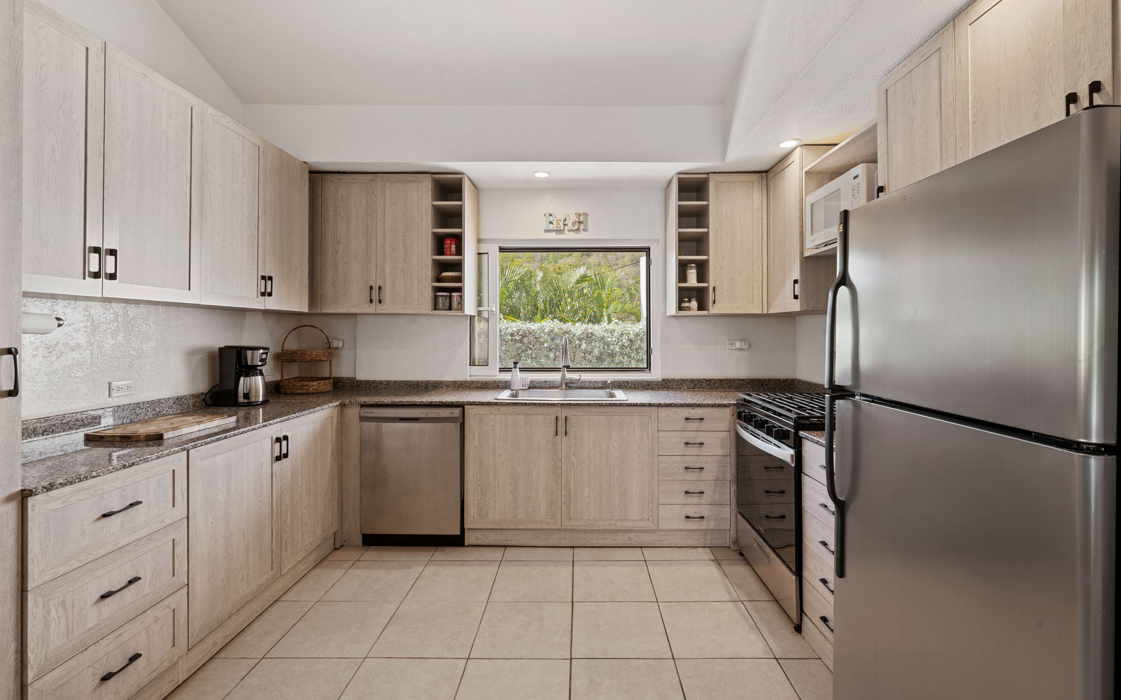 A modern kitchen with light wood cabinets, a stainless steel refrigerator, a built-in oven, and a window with a view of greenery.