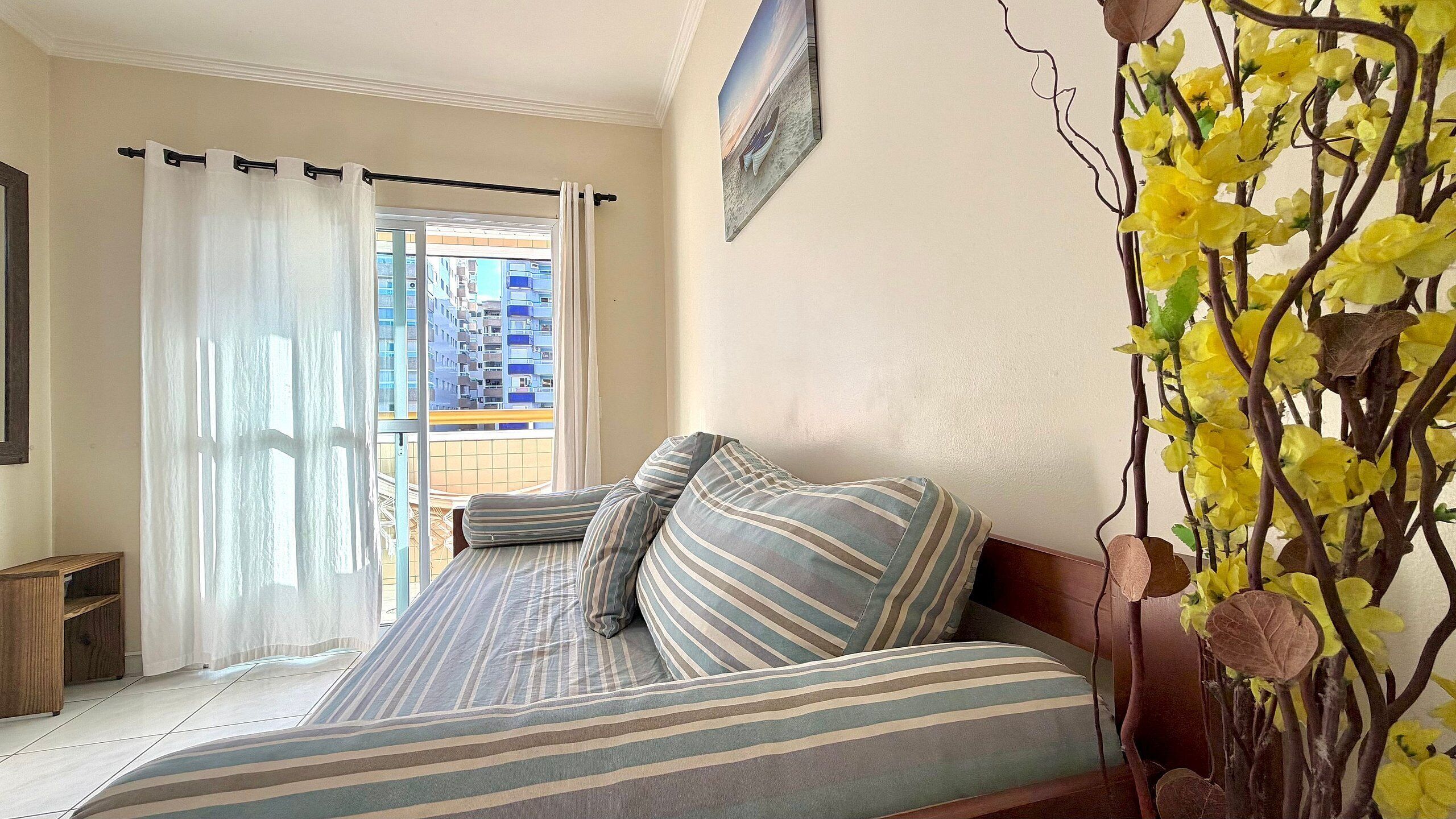 Una camera da letto con un letto, una finestra con tende, vista su edifici e un vaso con fiori.