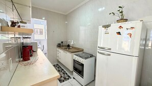 A compact kitchen with white appliances, a gas stove, a refrigerator with magnets, and a window with a view. - Residencial Texas (Praia Grande)