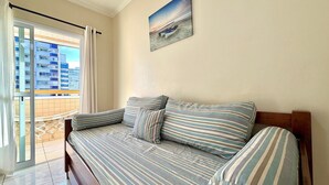 A room with a sofa bed, a view of buildings through a window, and a painting on the wall. - Residencial Texas (Praia Grande)
