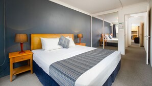 Ett hotellrum med en stor säng, ett nattduksbord med en lampa, en spegel på väggen och en dörr som leder till ett annat rum.