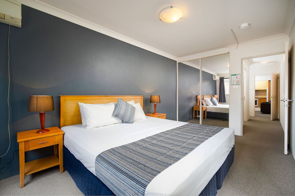 A hotel room with a large bed, a nightstand with a lamp, a mirror on the wall, and a door leading to another room.