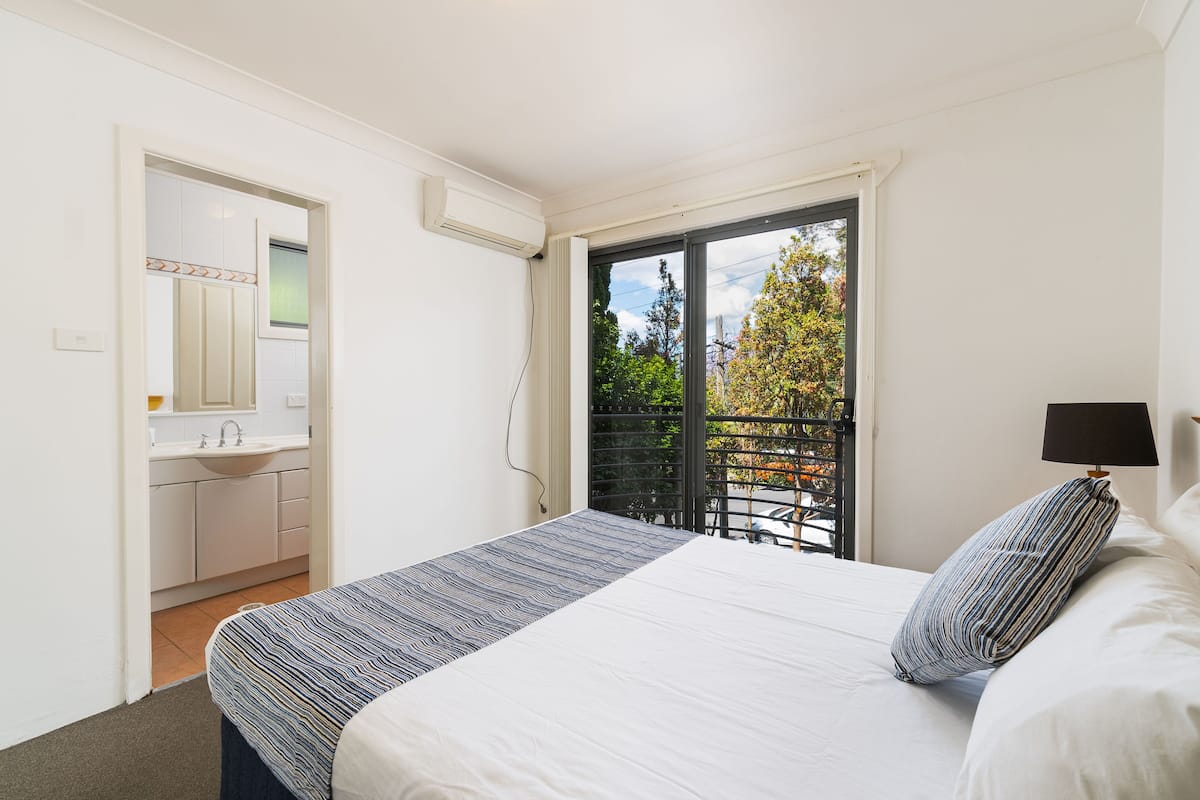 A bedroom with a bed, a nightstand, a window with a view of trees, and a bathroom with a sink and mirror.