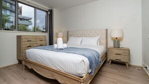 Une chambre à coucher avec un grand lit, une tête de lit en bois, deux tables de chevet et une fenêtre donnant sur des immeubles.