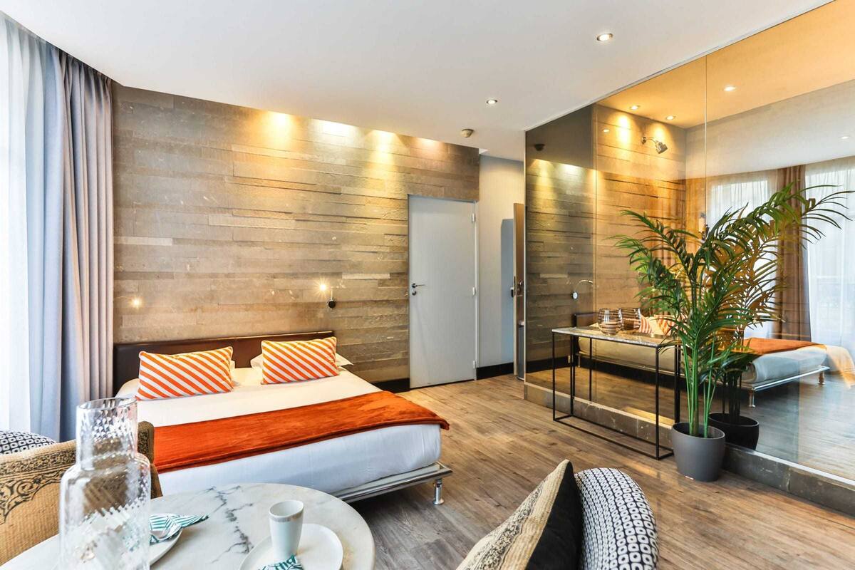 A modern hotel room with a large bed, a dining table, a sofa, and a potted plant.