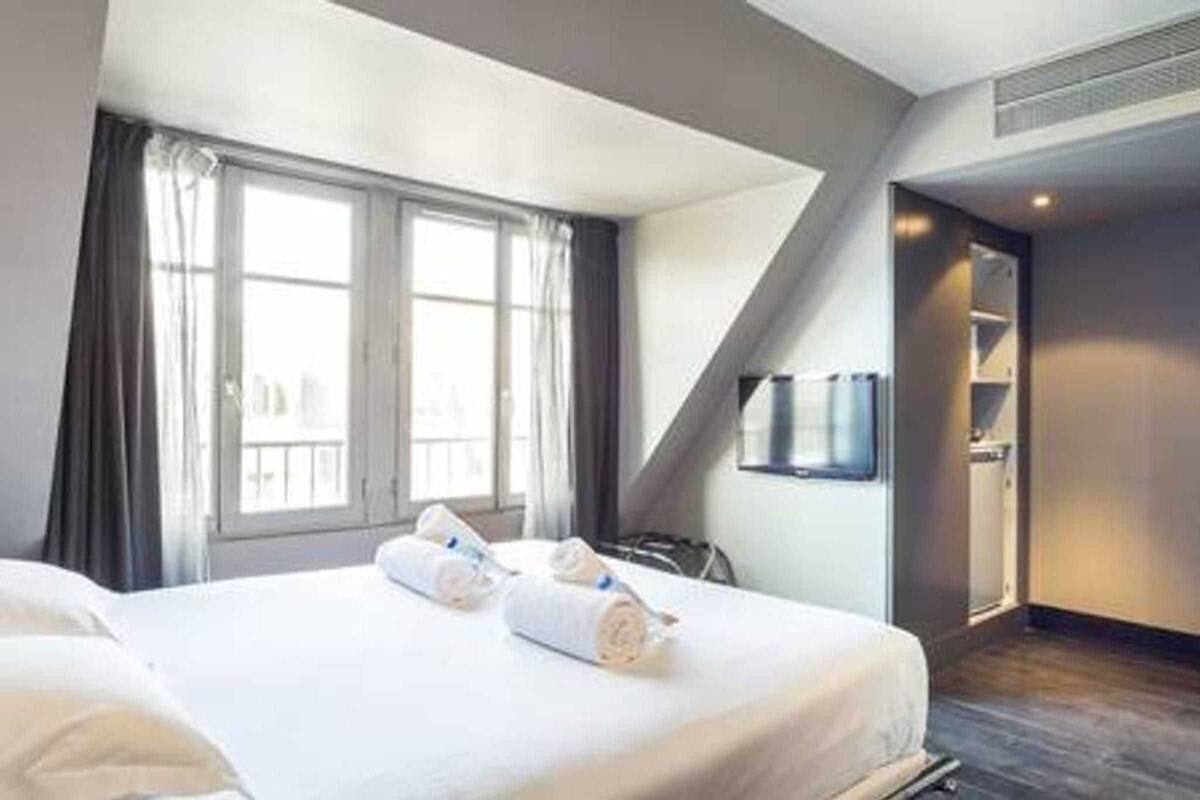A modern hotel room with a large bed, a TV mounted on the wall, and a view of the city through the window.