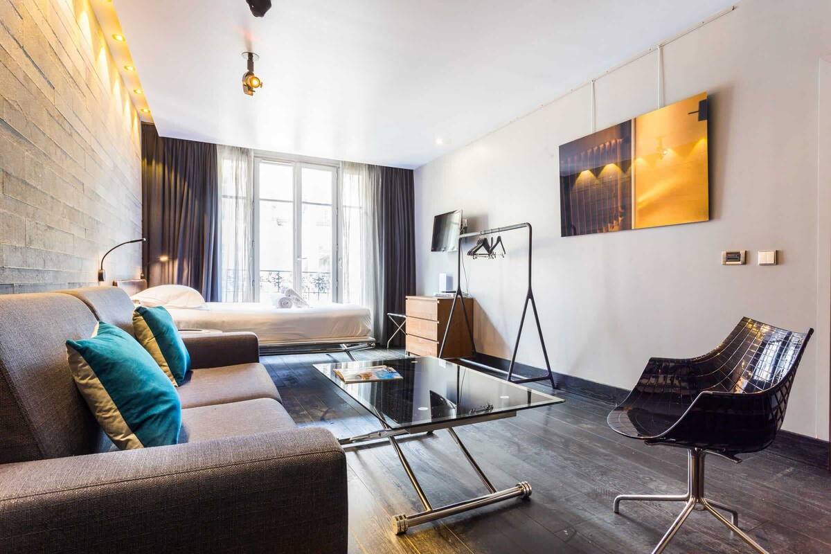 A modern hotel room with a sofa, a glass coffee table, a black chair, a TV, and a painting on the wall.