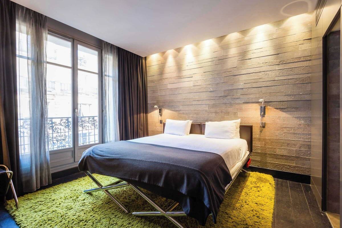 A modern hotel room with a large bed, a green carpet, a wooden accent wall, and a window with curtains.