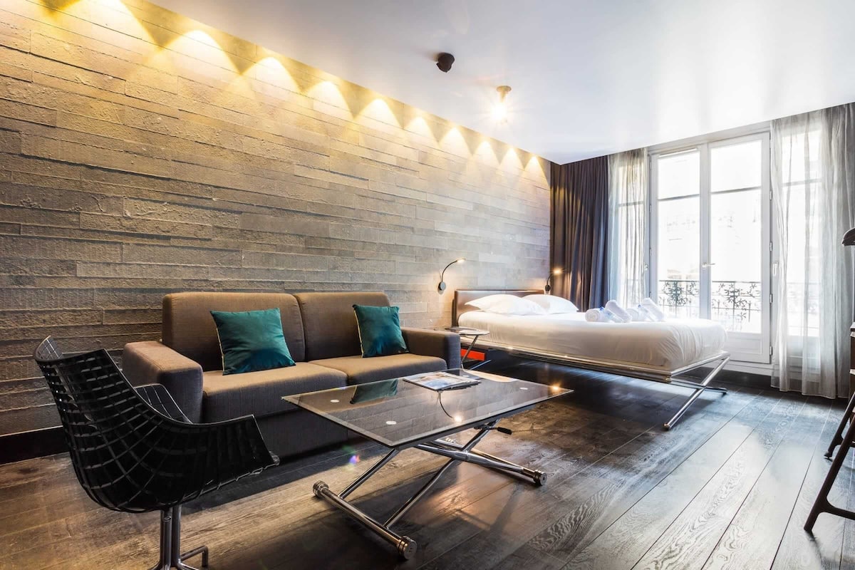 A modern hotel room with a sofa, a glass coffee table, a bed, and a view of the outdoors.