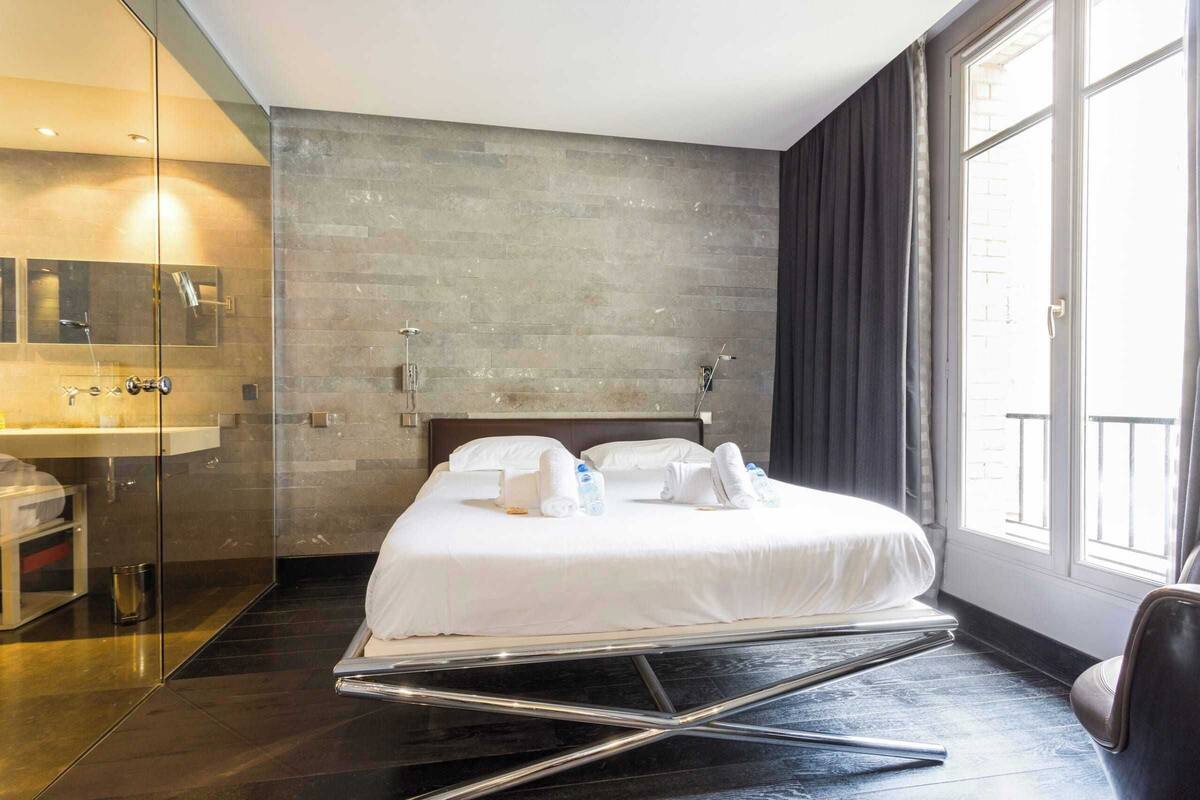 A modern hotel room with a large bed, a glass shower enclosure, a visible bathroom, and a view through a large window.