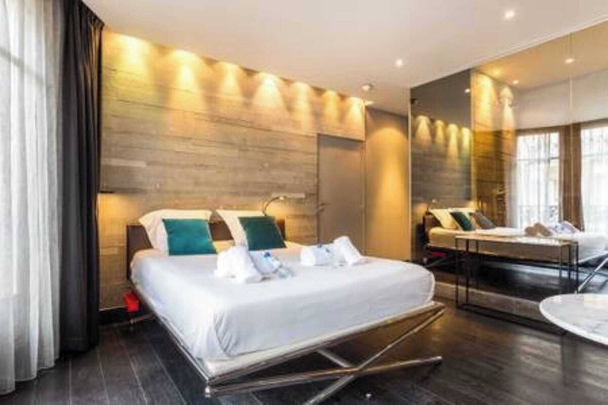 A modern hotel room with a bed, bedside tables, a mirror, and a view of the city.