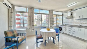 A modern kitchen with white cabinets, a dining area with a round table and blue chairs, and a view of buildings through large windows. - TERRASOL CARACOLA 2 (Torre del Mar)