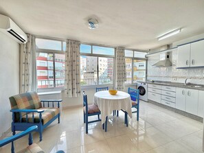 A modern kitchen with white cabinets, a dining area with a round table and blue chairs, and a view of buildings through large windows. - TERRASOL CARACOLA 2 (Torre del Mar)