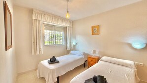 A room with two beds, a window with curtains, a bedside table with a lamp, and a wall-mounted light fixture. - TERRASOL CARACOLA 2 (Torre del Mar)