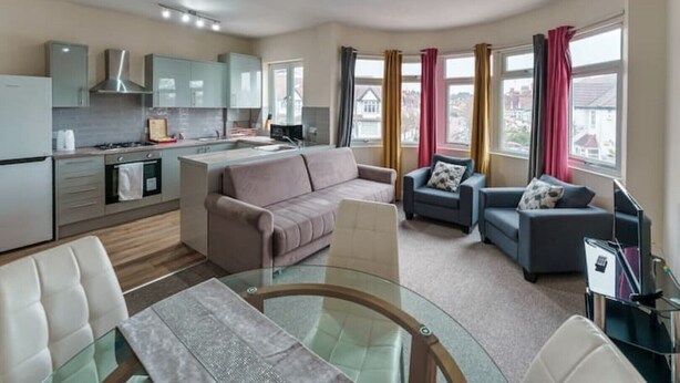A modern apartment with a kitchen, dining area, and living room. It features a glass dining table, a sofa, and a TV. - Fantasy accommodation in Golders Green/ Brent Cross  (London)