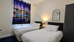 A hotel room with two single beds, a nightstand with a lamp, a window with curtains, and a wall-mounted clock. - Fantasy accommodation in Golders Green/ Brent Cross  (London)