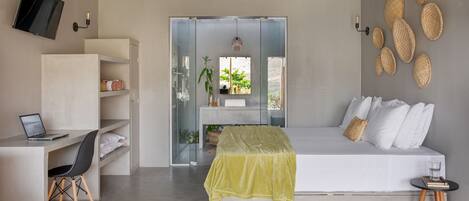 A modern bedroom with a bed, a desk with a laptop, and a view through a glass door to a bathroom.
