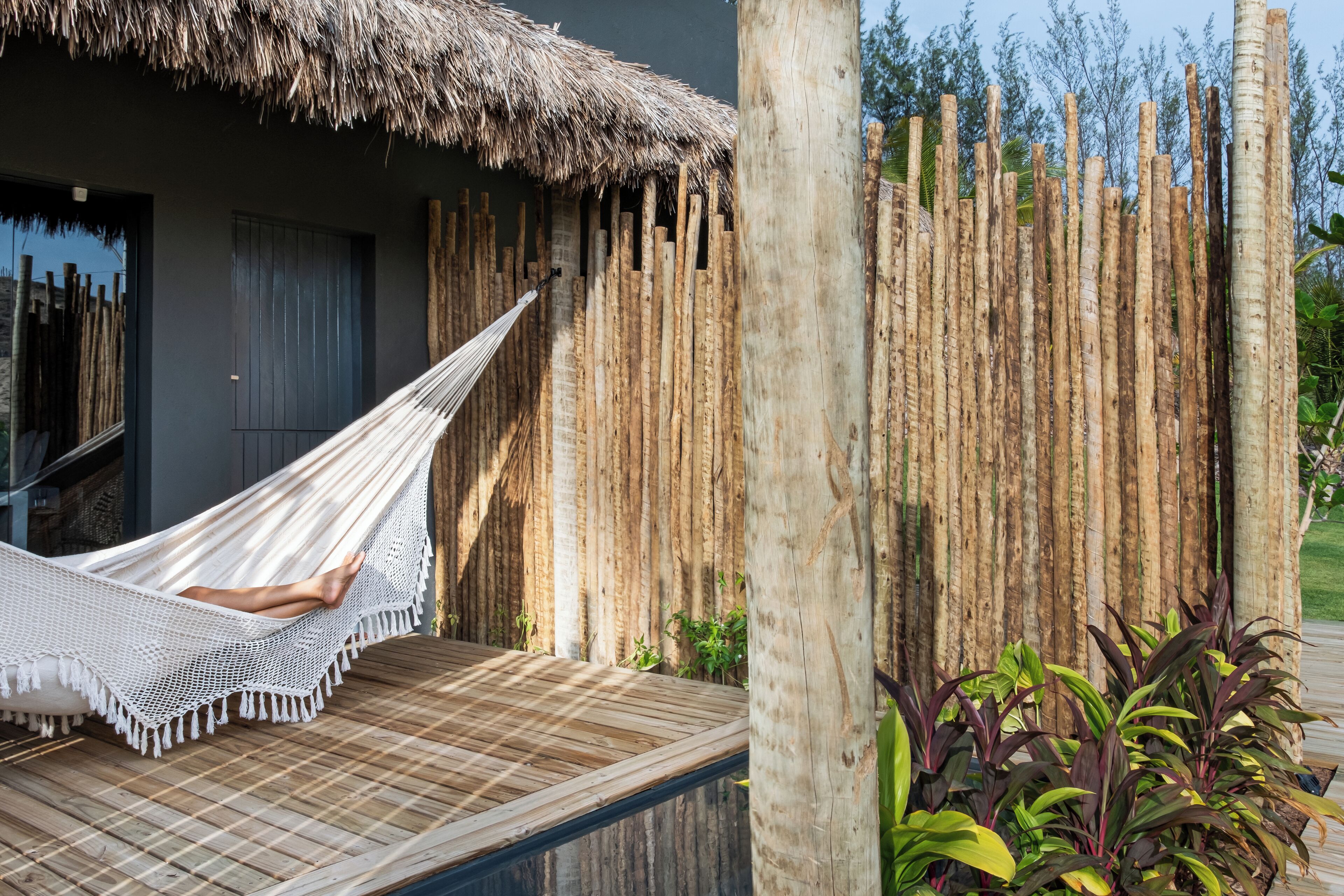 A hammock on a wooden deck, a thatched-roof structure, and a bamboo fence with plants.